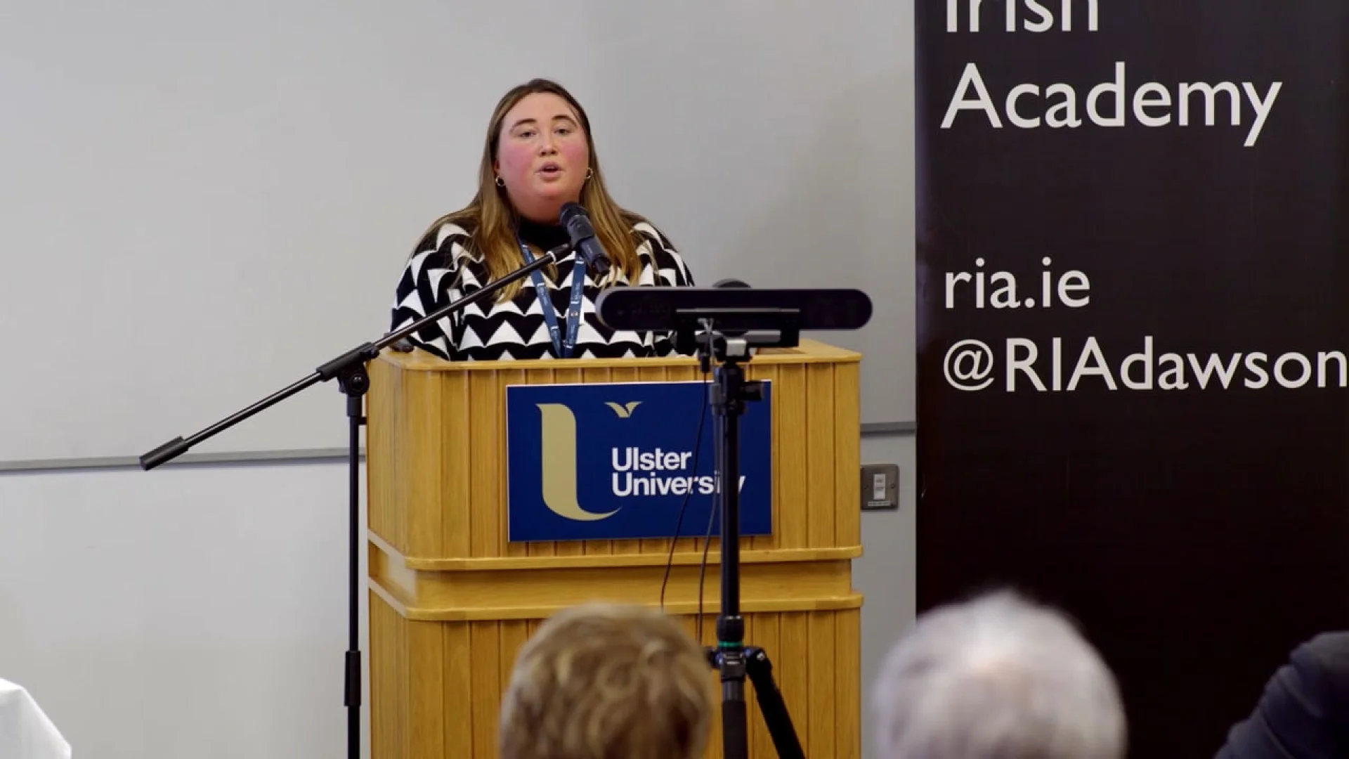 A speaker at a podium during Rhyming Weavers Anniversary: Part Four, showcasing brand development strategy at an Ulster University event.