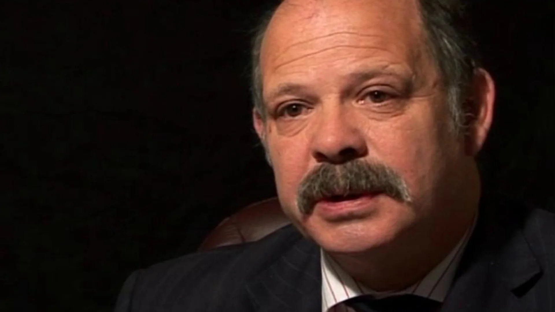 Close-up of a middle-aged man with a gray mustache wearing a dark suit and striped shirt, against a dark background.