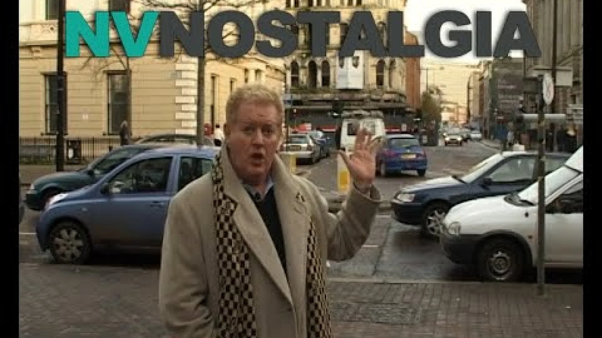 A man in a beige coat and patterned scarf stands on a city street, gesturing with his hand in front of passing cars.