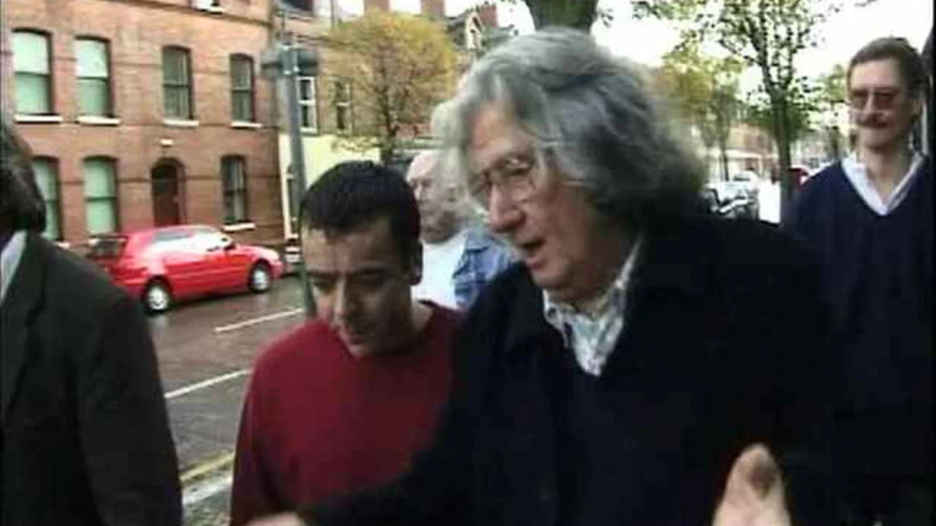 Older man with gray hair in a dark sweater gestures as he walks beside younger men along a city street with red brick buildings and a red car in the background