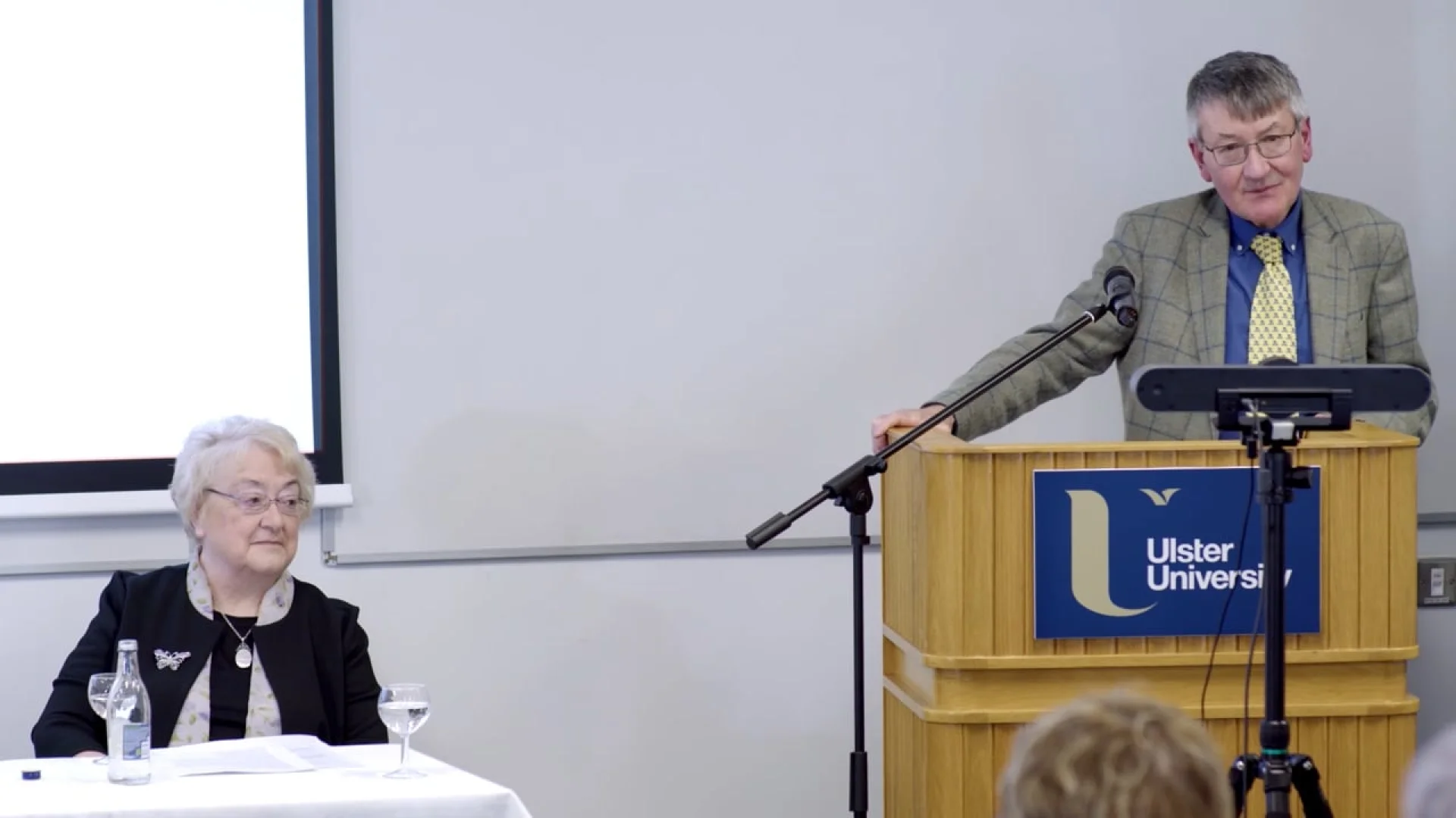 A speaker at an Ulster University podium for Rhyming Weavers Anniversary: Part Two, with an audience member seated nearby.