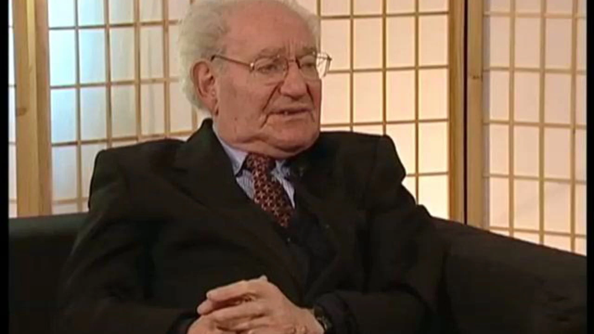 Older man with gray hair and glasses in a dark suit and patterned tie sits with hands clasped during an interview, wooden grid backdrop behind him