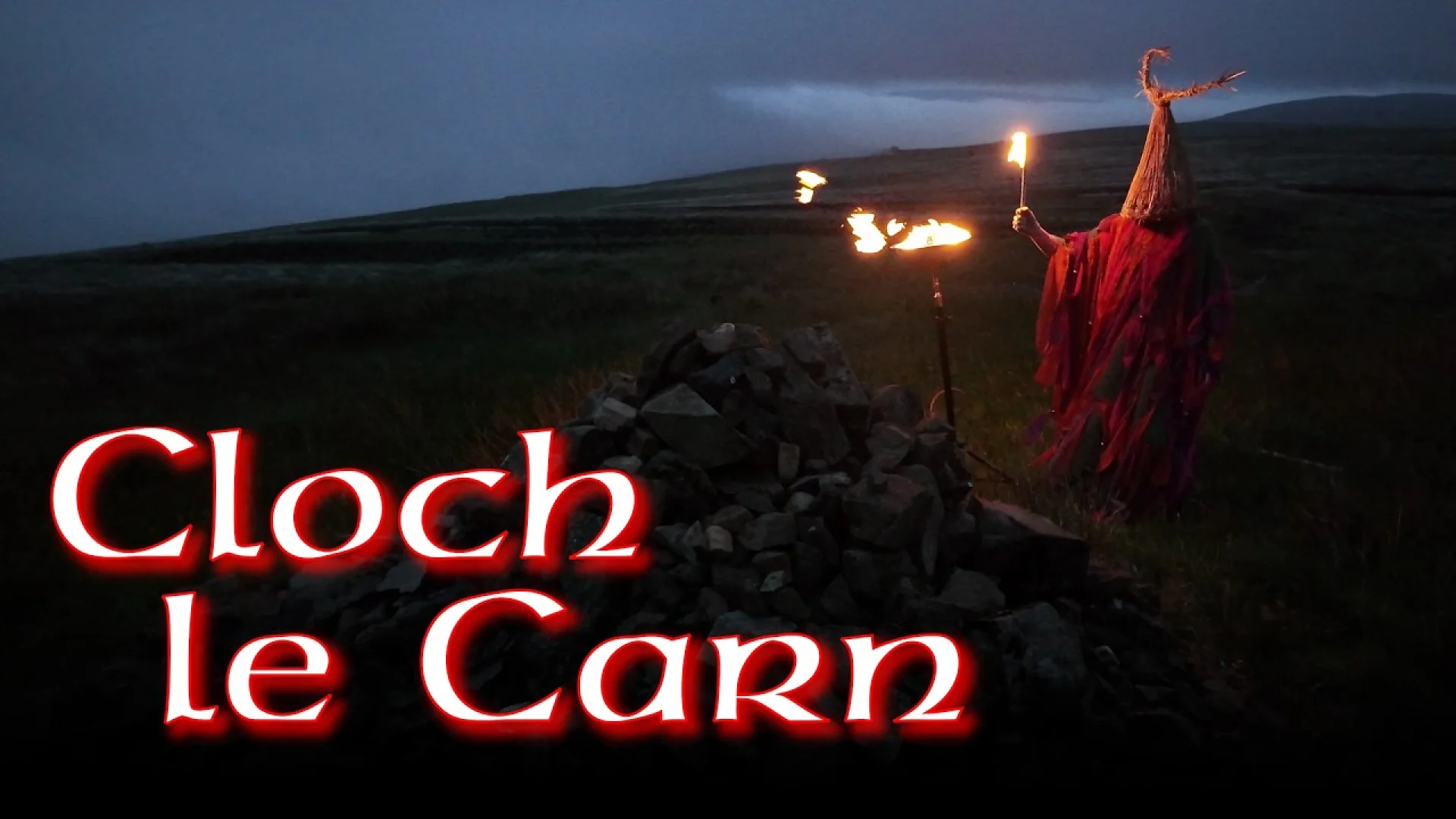 A figure in a straw mask and red cloak holds a torch by a stone cairn, highlighting Cloch le Carn – Tógáil Pobail Charn Tóchair brand strategy.
